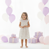 Girl wearing white floral smocked bishop dress with purple balloons and gift boxes