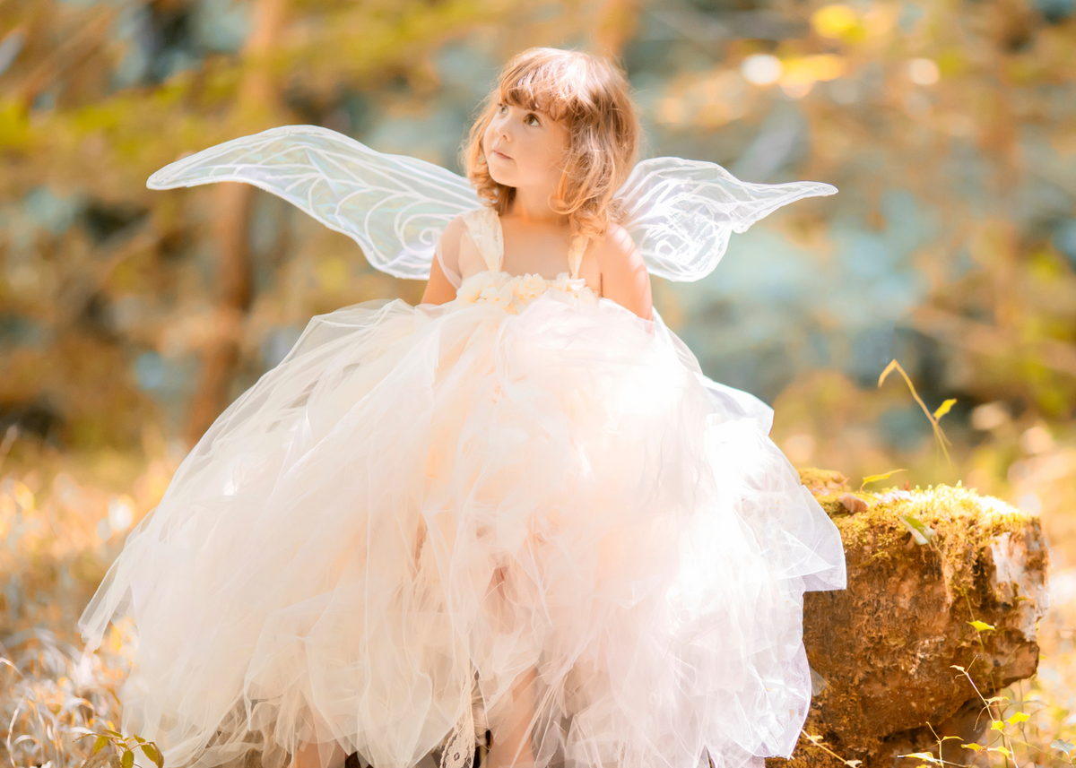 A little girl with wavy hair gazes upward beside a moss-covered tree stump in a sunlit autumn woodland. She wears an ivory fairy dress with a full, layered tulle skirt, and translucent wings, surrounded by golden foliage and light filtering through trees.