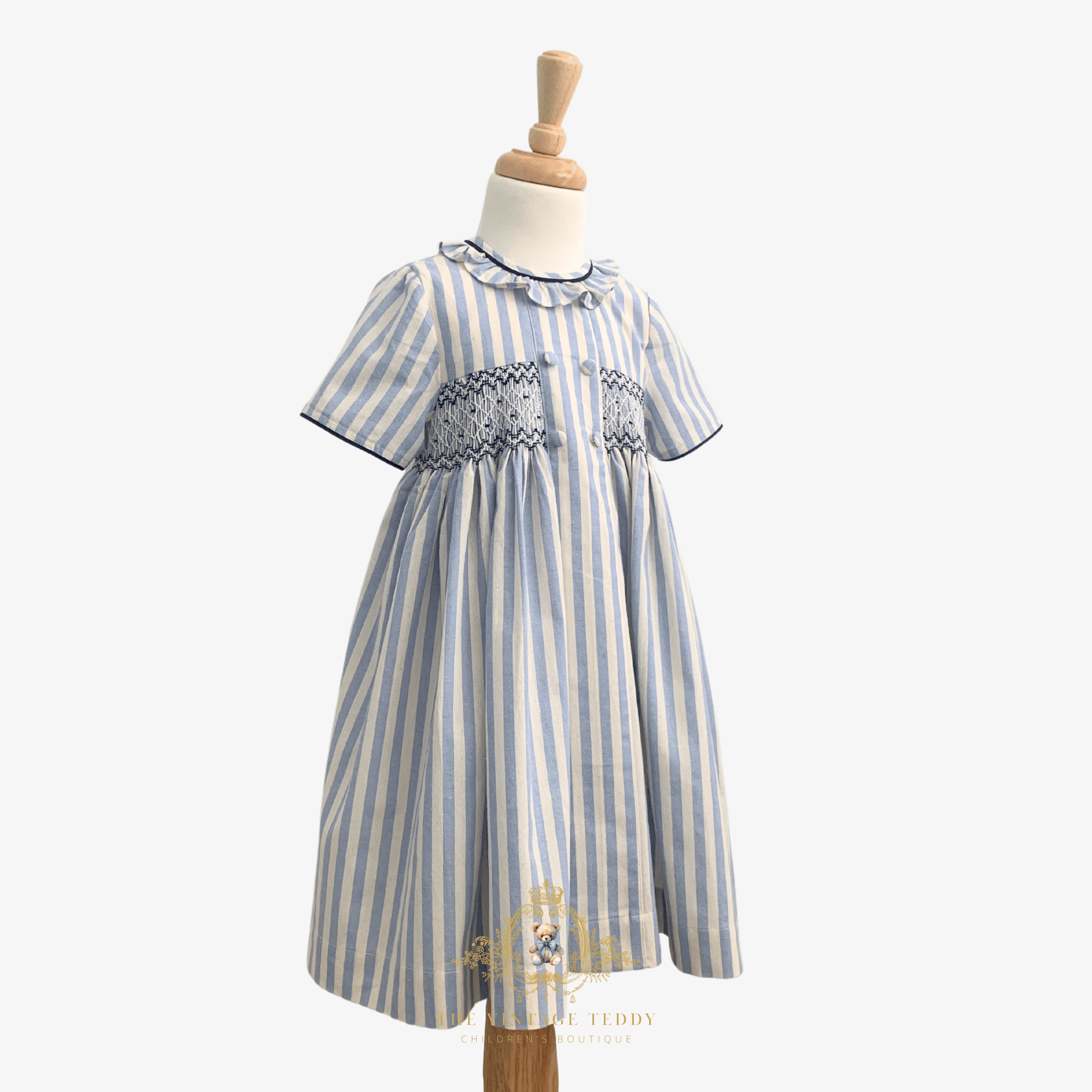 A short-sleeve toddler girls dress with vertical blue and cream stripes and a smocked chest area, displayed on a wooden mannequin dress form against a light white background.