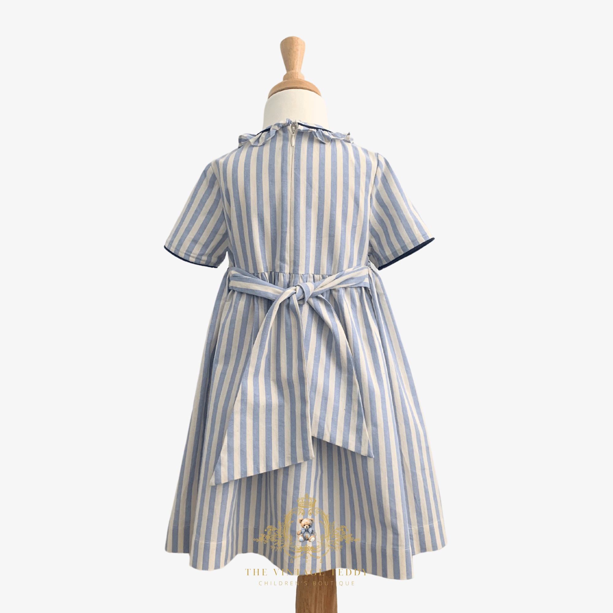 Back of a short-sleeve child's dress with vertical light blue and white stripes, displayed on a mannequin. The dress features a ruffled collar, a bow that ties at the waist, and a layered asymmetrical hem.
