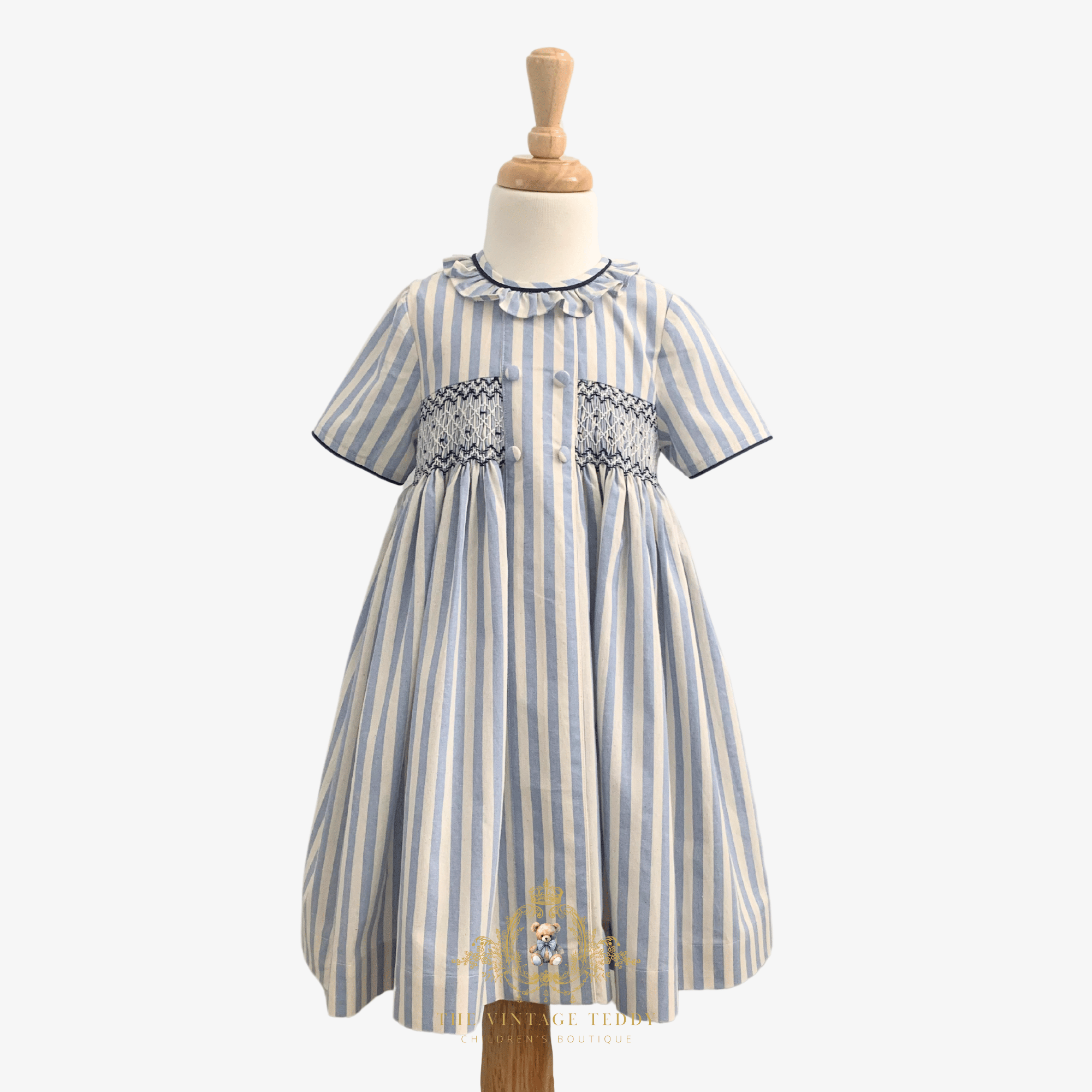 A short-sleeve baby girl's dress with vertical light blue and cream stripes, featuring a smocked chest and ruffled collar, displayed on a wooden dress form mannequin against a light background.