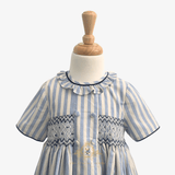 A baby or toddler girl's short-sleeve dress with vertical light blue and cream stripes, featuring a ruffled collar, decorative smocked chest panel, and front buttons, displayed on a white dress form with a wooden neck cap.