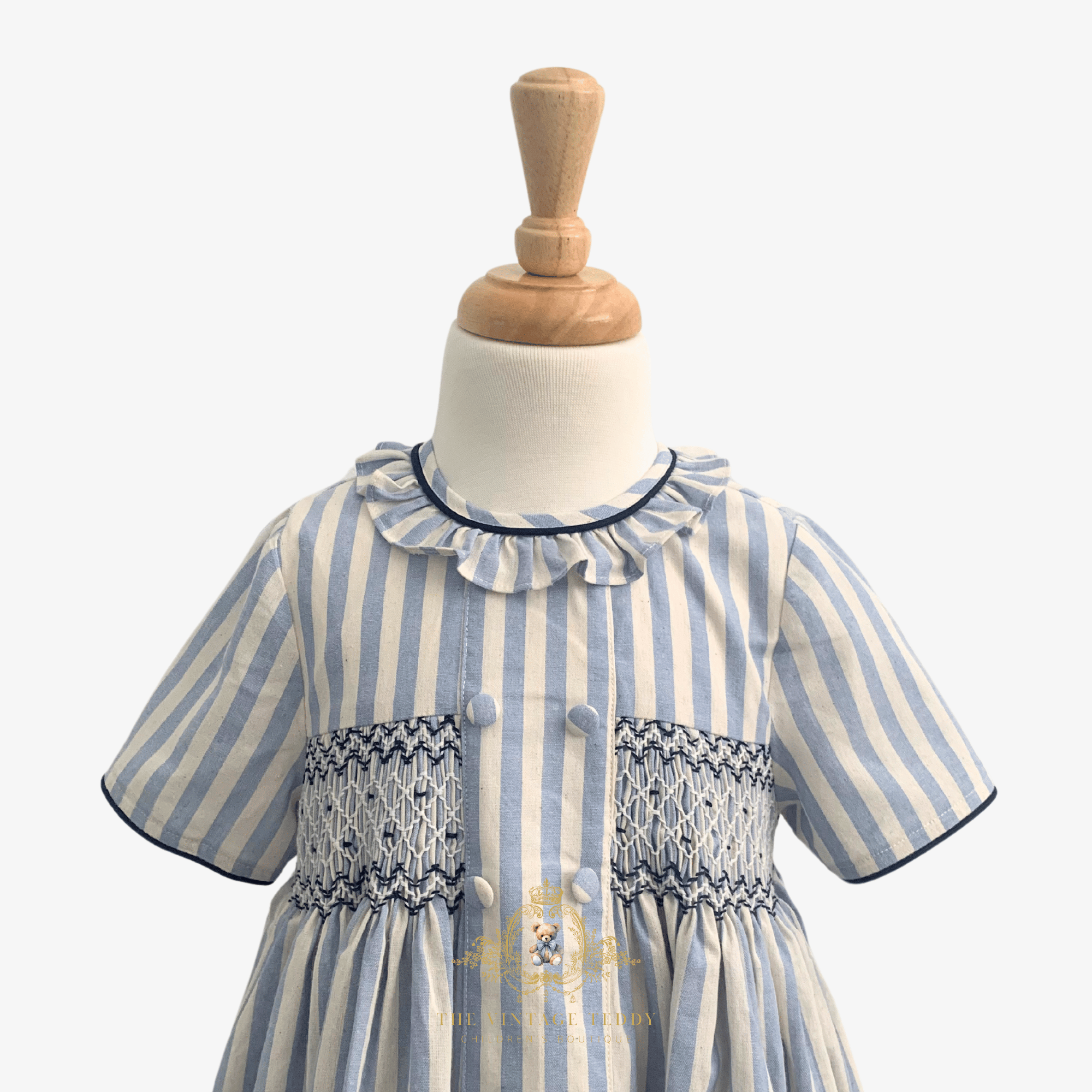 A baby or toddler girl's short-sleeve dress with vertical light blue and cream stripes, featuring a ruffled collar, decorative smocked chest panel, and front buttons, displayed on a white dress form with a wooden neck cap.