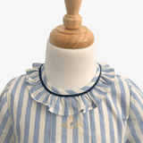 A close-up of a child's blue and cream striped kids dress with a ruffled collar trimmed in navy blue, displayed on a white mannequin torso with a wooden neck cap.