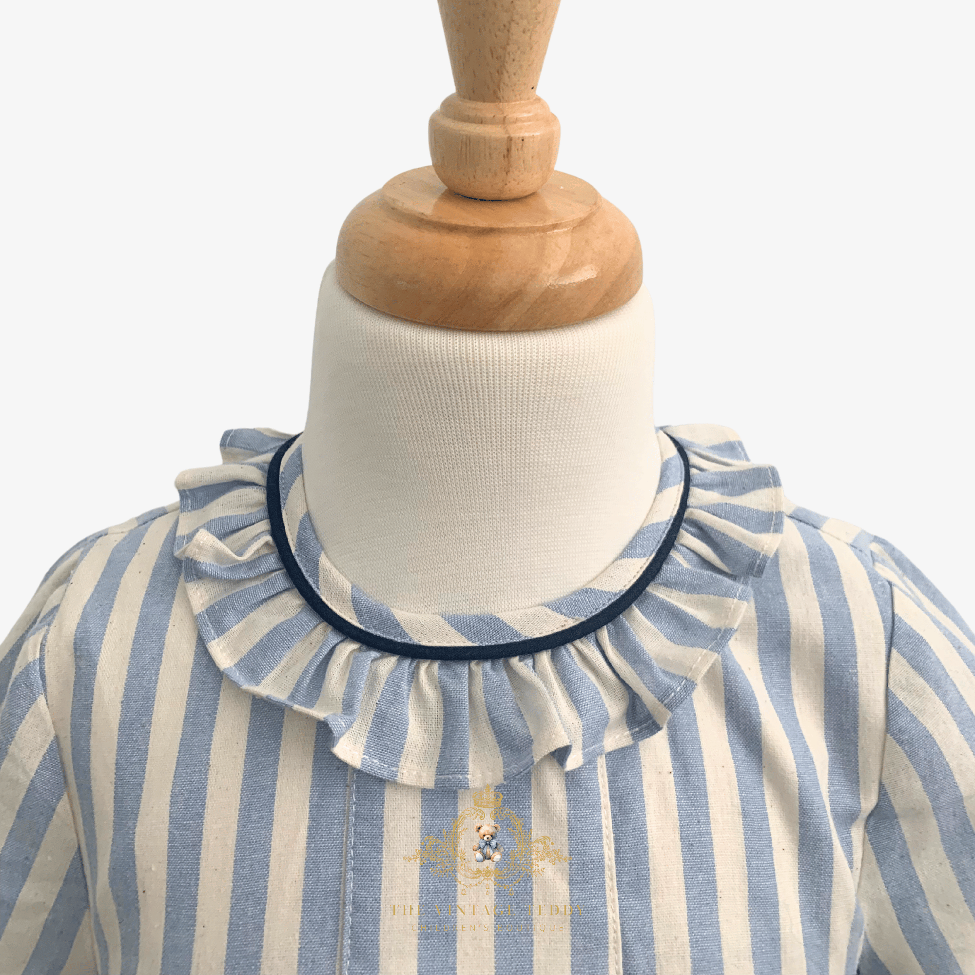 A close-up of a child's blue and cream striped kids dress with a ruffled collar trimmed in navy blue, displayed on a white mannequin torso with a wooden neck cap.