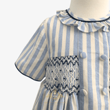 A close-up of a child's dress sleeve on a mannequin, featuring vertical light blue and cream stripes, a ruffled collar with dark blue trim, and a smocked chest panel with decorative buttons.