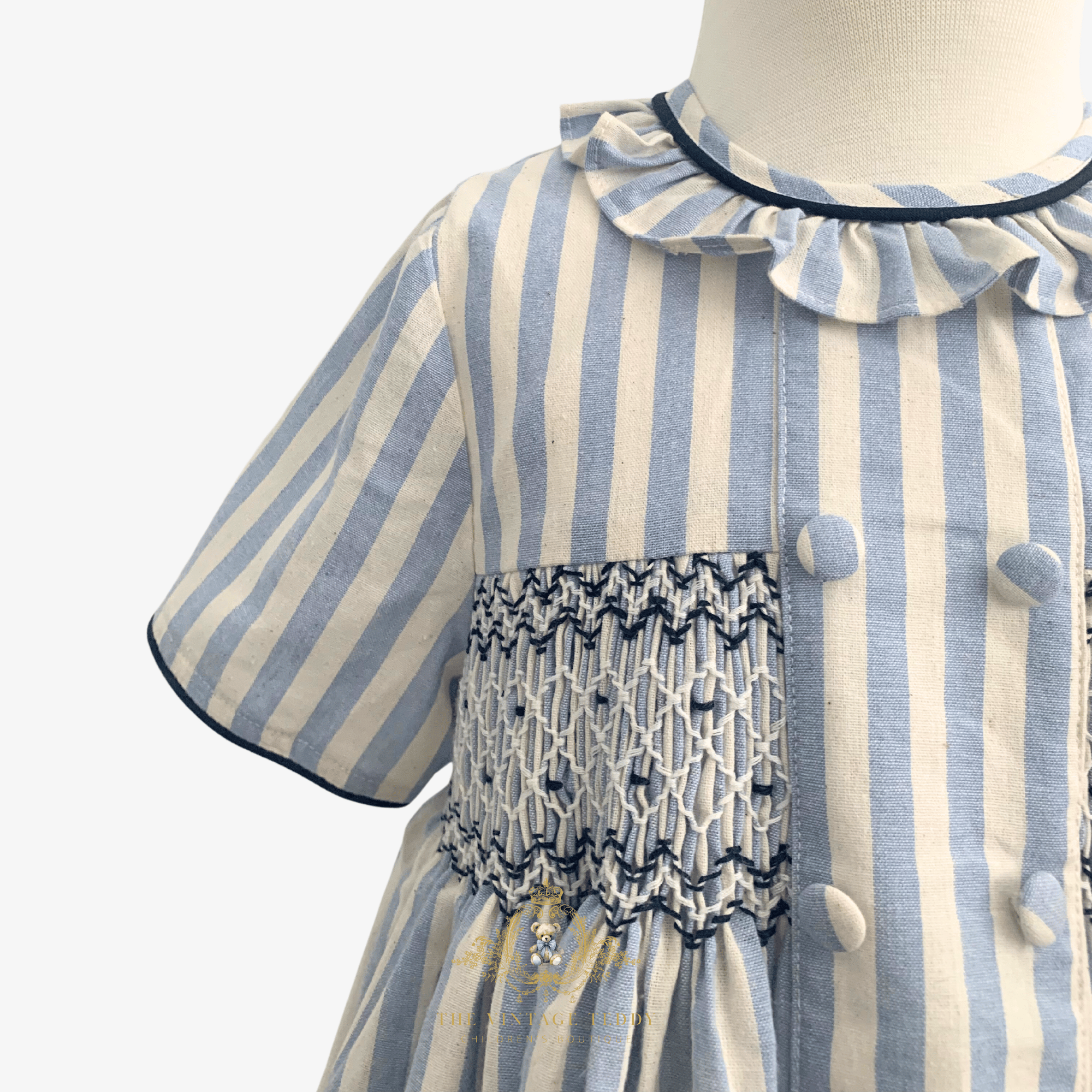 A close-up of a child's dress sleeve on a mannequin, featuring vertical light blue and cream stripes, a ruffled collar with dark blue trim, and a smocked chest panel with decorative buttons.