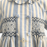 Close-up of a vintage-style striped children's smocked dress with vertical cream and dusty blue stripes, decorative smocked panels with navy blue embroidery, and matching blue buttons.