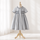 A short-sleeve girl's dress with vertical blue and white stripes, featuring smocked detailing across the chest and a ruffled collar, displayed on a wooden dress form mannequin in a bright, softly lit room.