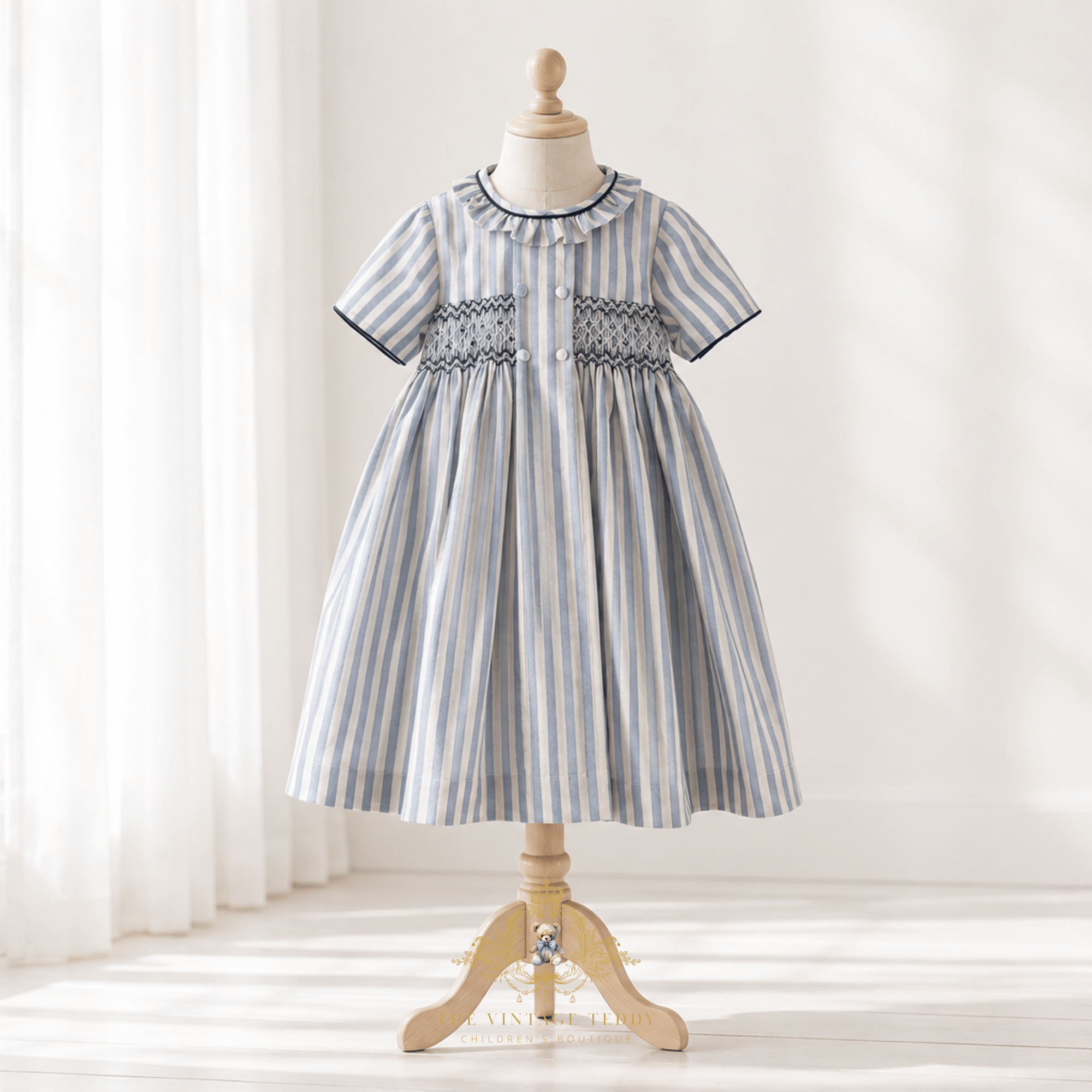 A short-sleeve girl's dress with vertical blue and white stripes, featuring smocked detailing across the chest and a ruffled collar, displayed on a wooden dress form mannequin in a bright, softly lit room.