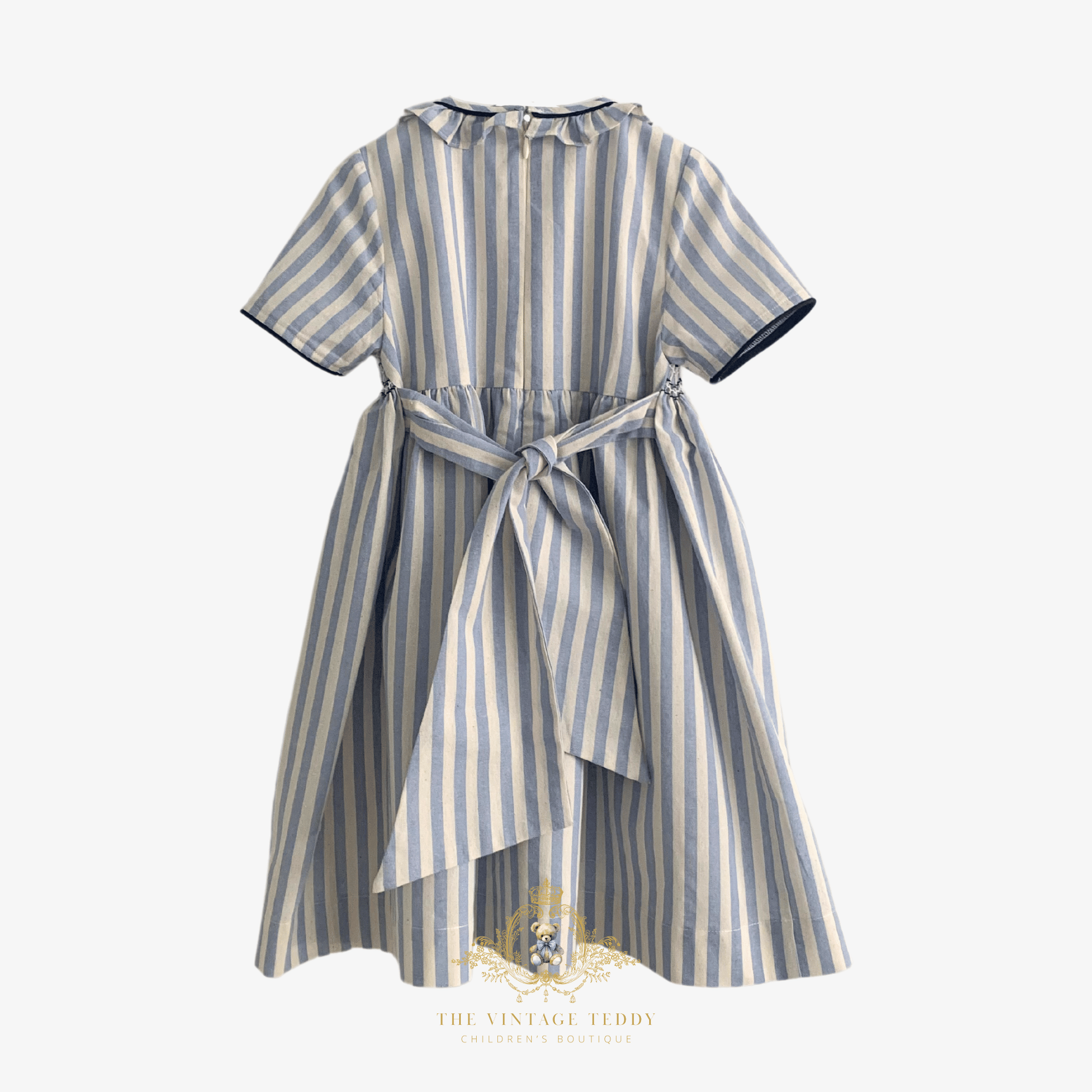 The back of a short-sleeve children's dress with vertical blue and beige stripes, featuring a bow at the back and a ruffled collar, shown against a light background.