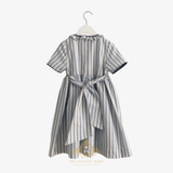 The back of a short-sleeve children's dress with vertical blue and cream stripes, featuring a ruffled collar, bow, and a knee-length dress skirt, shown hanging on a white hanger against a light background.