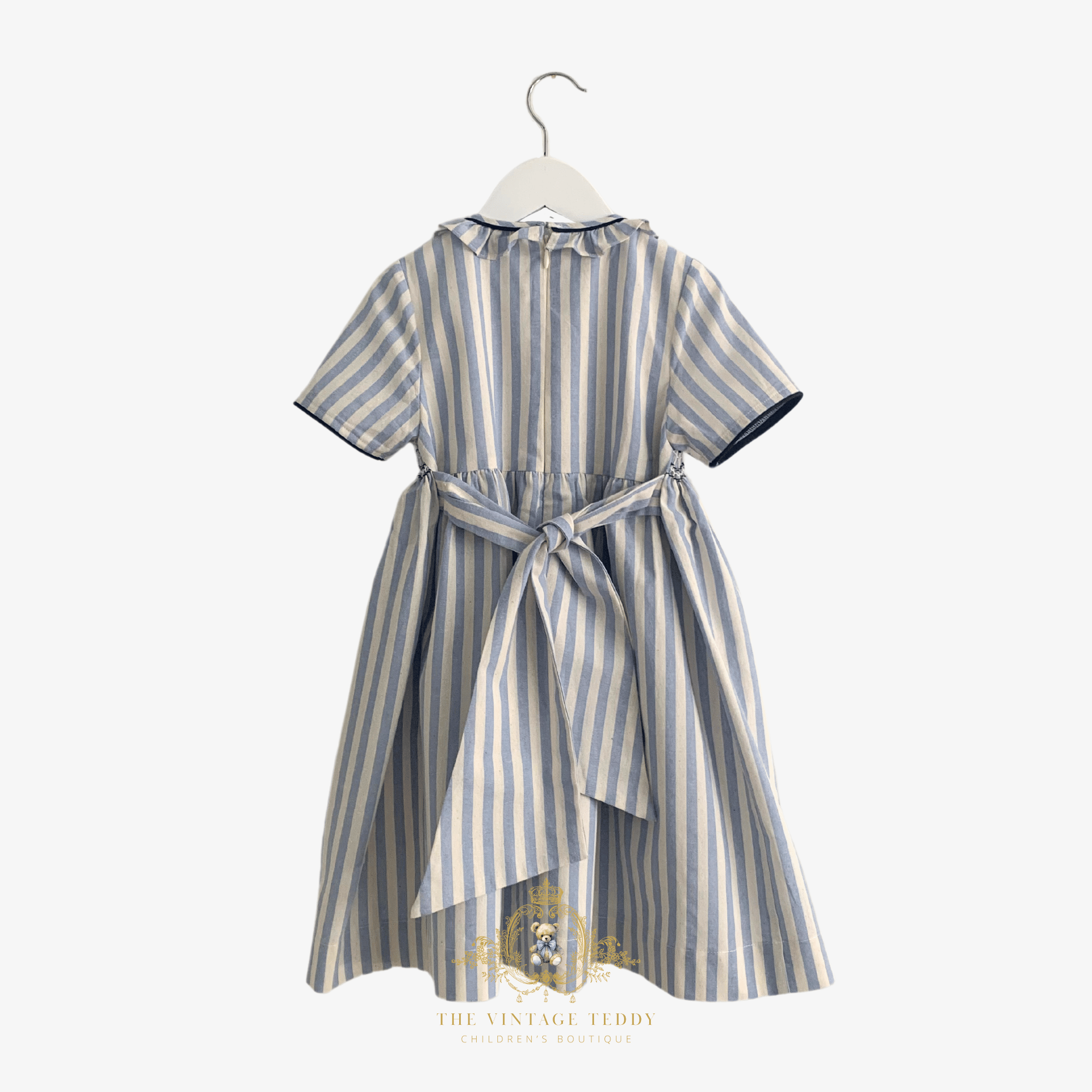 The back of a short-sleeve children's dress with vertical blue and cream stripes, featuring a ruffled collar, bow, and a knee-length dress skirt, shown hanging on a white hanger against a light background.