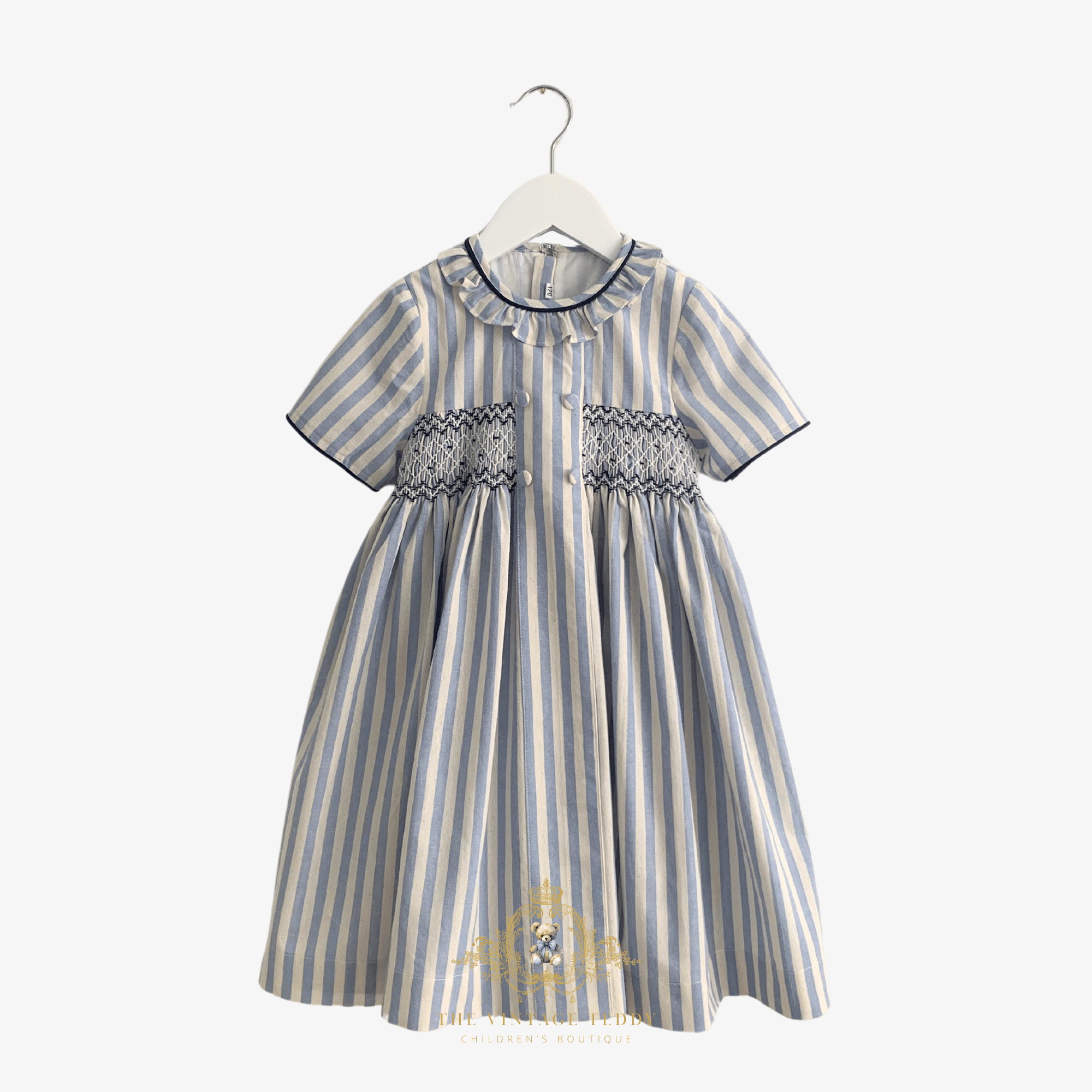 A short-sleeve baby or toddler girls dress with vertical blue and beige stripes, featuring a smocked bodice, hanging on a white hanger against a light  background.