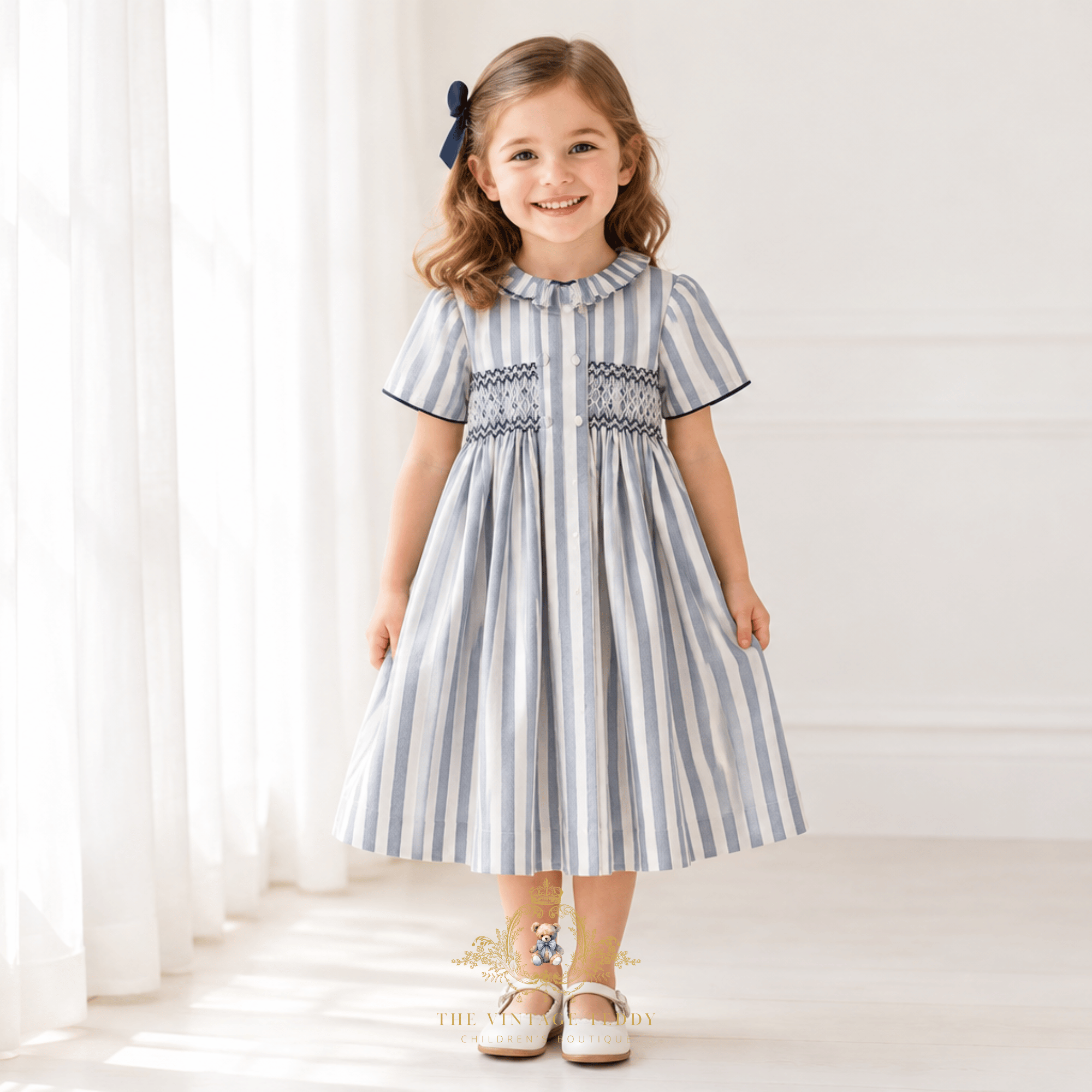 A young girl stands in a bright room wearing a short-sleeve dress with vertical blue and white stripes and a smocked bodice. She has a navy hair bow and is wearing a smile and white Mary Jane shoes.