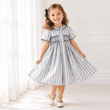 A young girl with a navy hair bow is wearing a short-sleeve, blue and white striped dress with smocked details on the chest. She is standing in a bright room with white curtains and light-colored walls, holding out her skirt to show its volume.