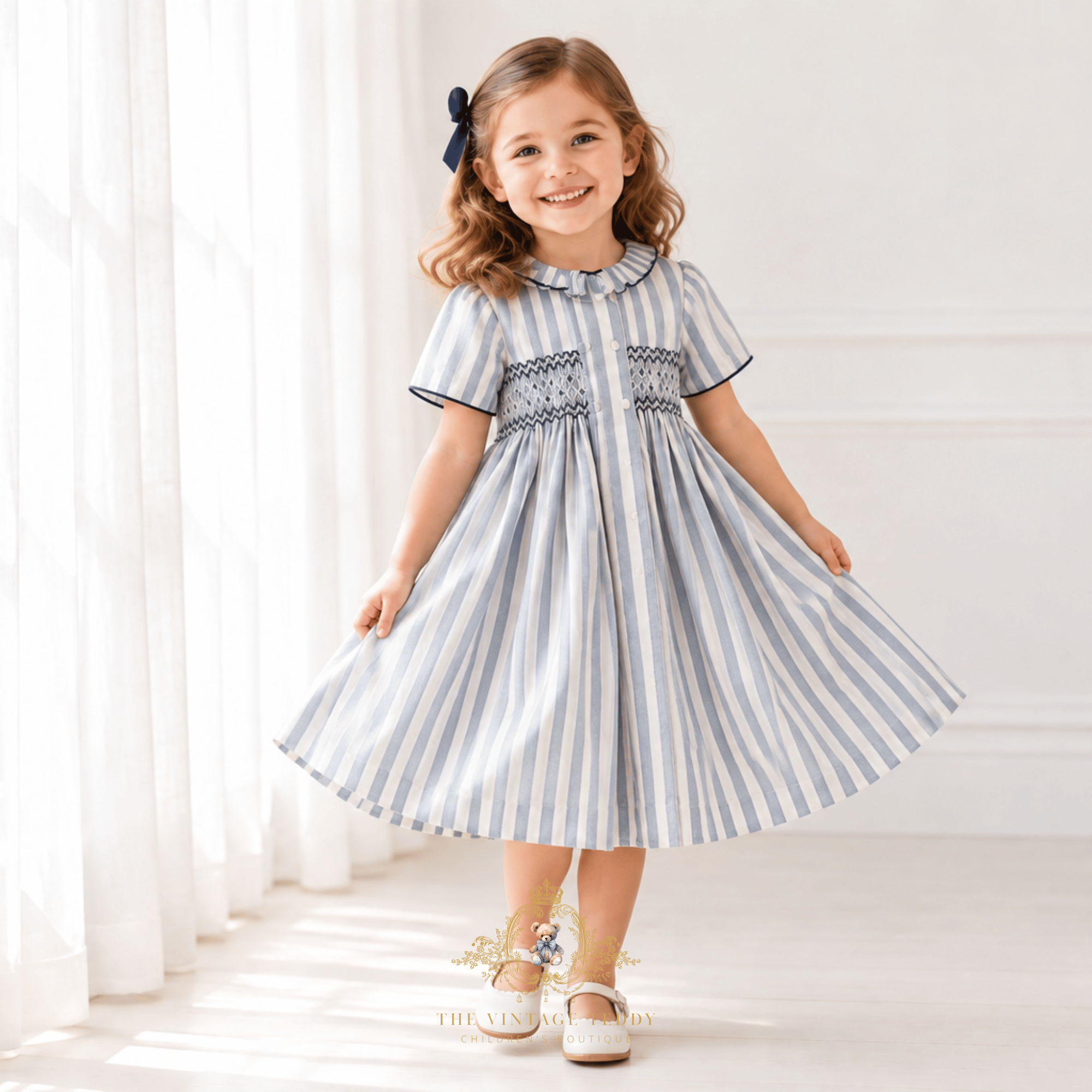 A young girl with a navy hair bow is wearing a short-sleeve, blue and white striped dress with smocked details on the chest. She is standing in a bright room with white curtains and light-colored walls, holding out her skirt to show its volume.