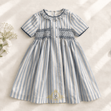 A short-sleeve baby or toddler girls dress with vertical light blue and white stripes, featuring a smocked bodice and ruffled collar, laid flat on a textured cream fabric background with white baby's breath flowers on the left.